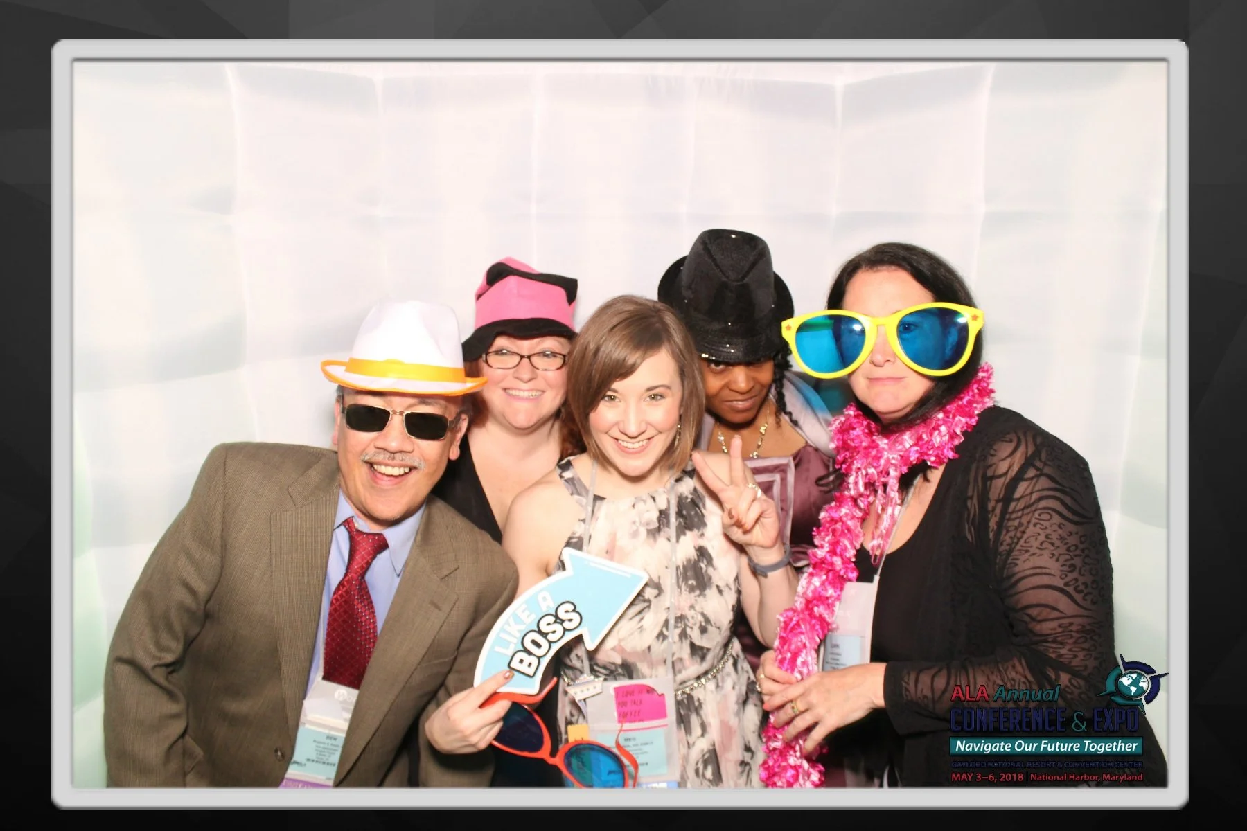 Group of five people at a conference photo booth, wearing colorful accessories like hats, sunglasses, and leis, smiling and posing for the camera. The banner at the bottom mentions the ALA Annual Conference & Expo held in May 2018 in Maryland.
