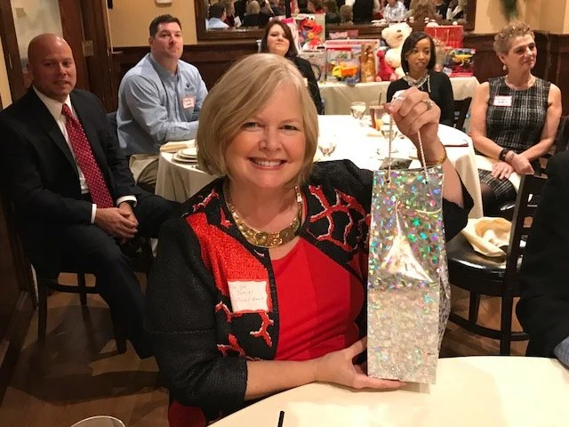 A woman with blonde hair smiling and wearing a red dress with a black and red patterned jacket is holding a glittery gift bag at a banquet or party. She is sitting at a table with other guests, some of whom are looking toward her. The background show