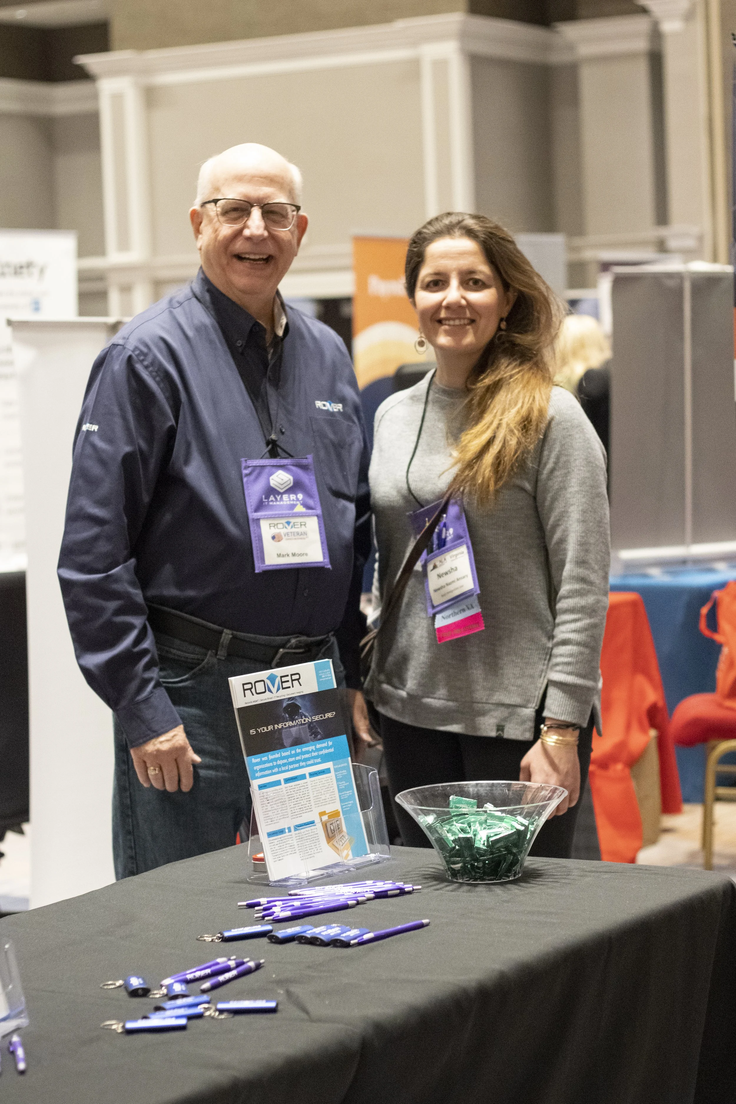 Two people, a man and a woman, standing behind a table at a conference or event. The table has promotional items, including pens and lanyards, and a brochure. Both are wearing badges and are smiling at the camera.