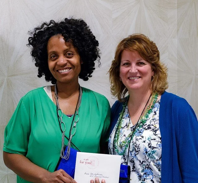 Two women standing side by side smiling, one holding a gift card and a card, against a light patterned wall.
