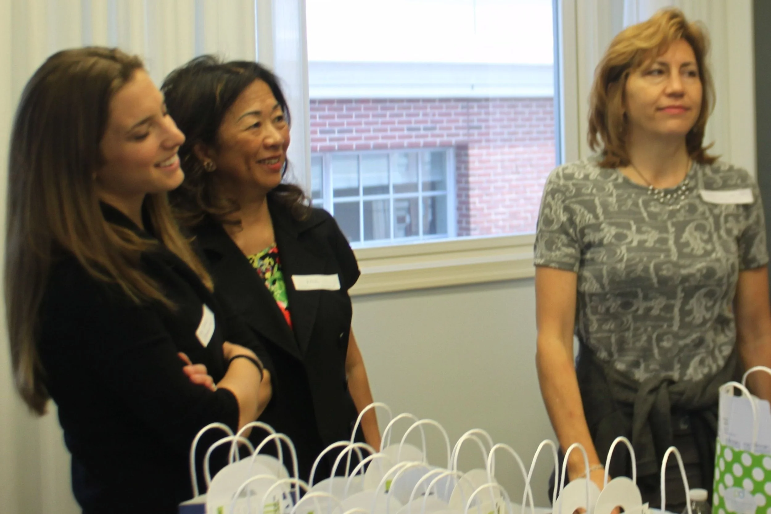Three women standing in a room near a window, smiling and observing white gift bags arranged on a table in front of them.