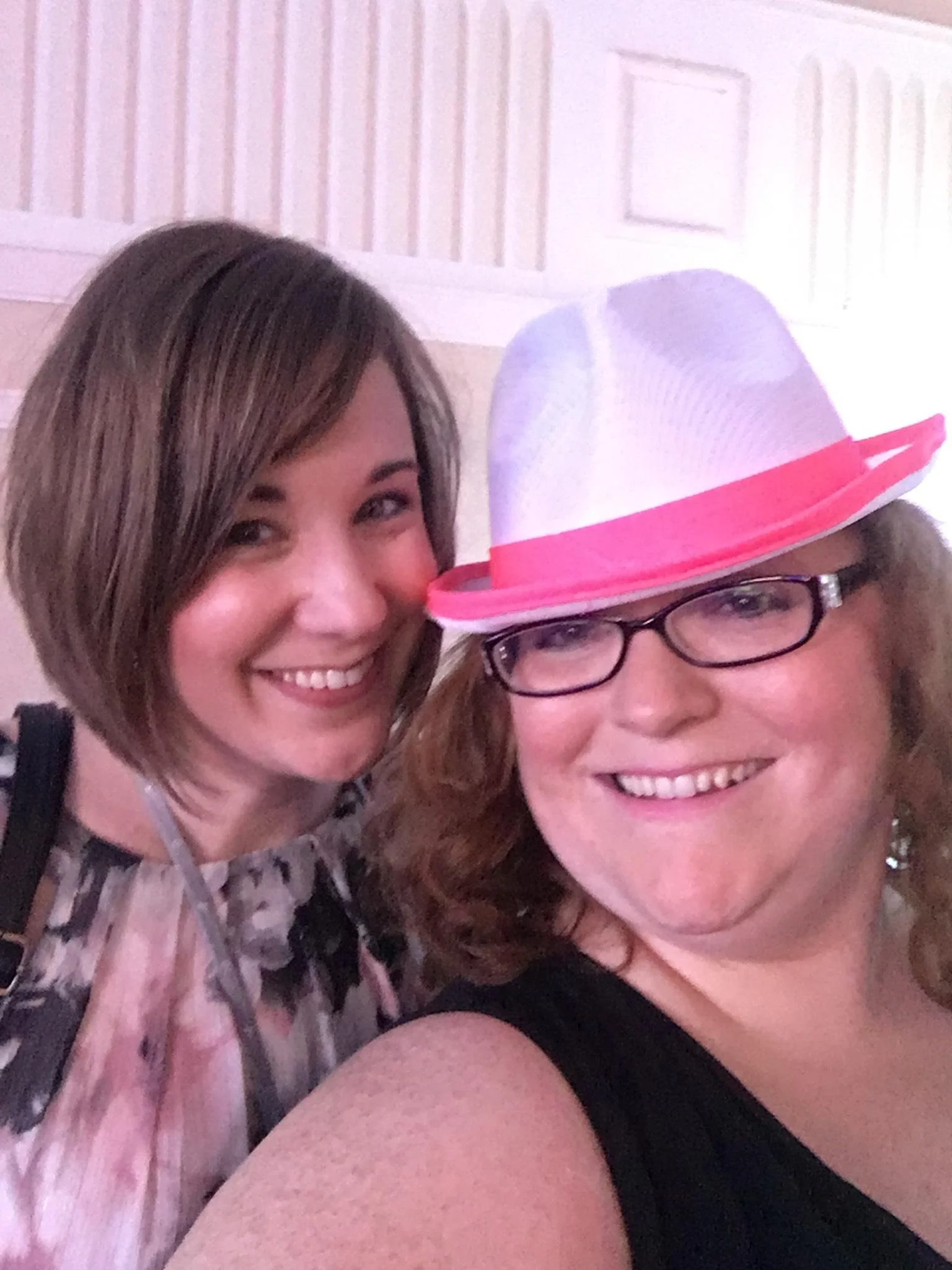 Two women smiling, one wearing glasses and a pink and purple hat, with a light-colored background.