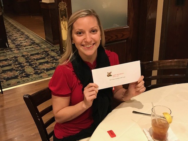 A woman sitting at a dining table, smiling, holding an envelope. There is a drink with a lemon slice and a straw on the table.