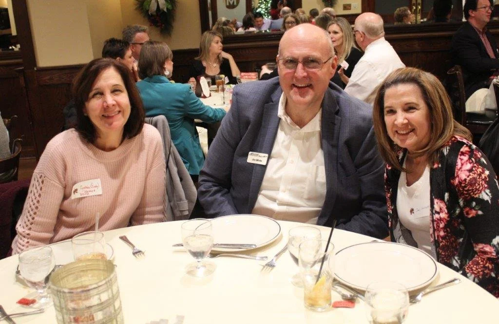 Three people sitting at a round table at a restaurant or banquet, with other people in the background. The person in the middle is a bald man with glasses, wearing a blazer, flanked by two women smiling at the camera.