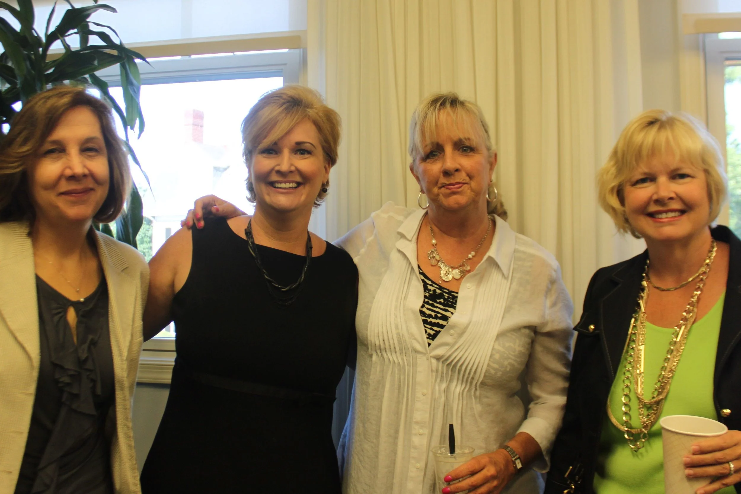 Four women standing together indoors, smiling and posing for a photo.