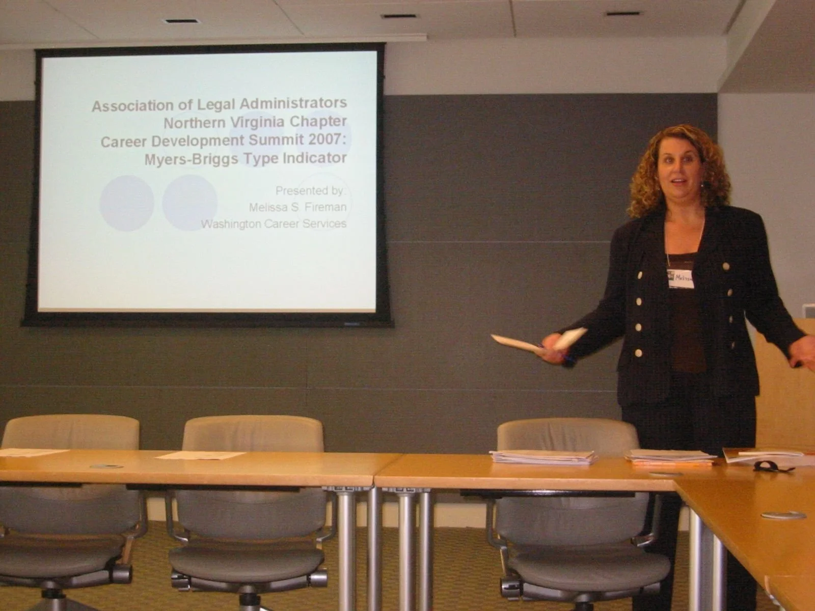 A woman in black attire standing in front of a screen giving a presentation at a career development summit, with chairs and a table in the foreground.