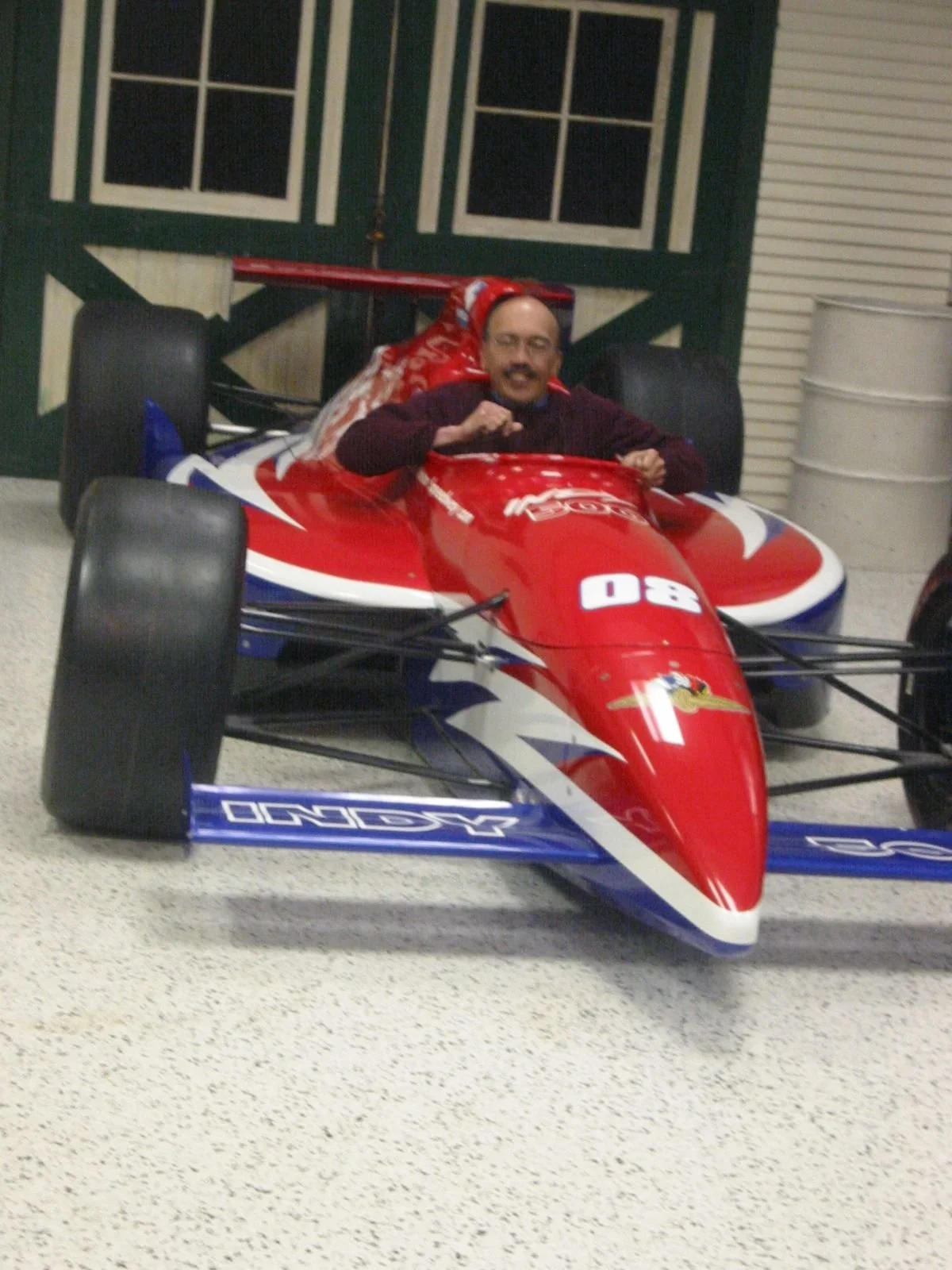 A man sitting in a red, white, and blue Formula race car with the number 80 on it, inside a garage with a closed green barn door and a white barrel