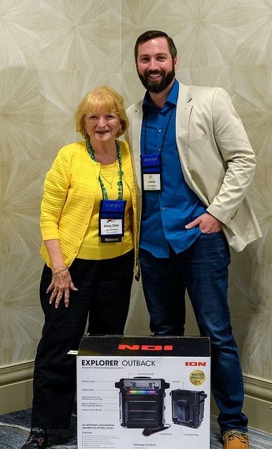 A woman and a man standing together indoors, smiling, with name badges around their necks. The woman is wearing a yellow jacket and the man is in a beige blazer and blue shirt. There is a product box in front of them.