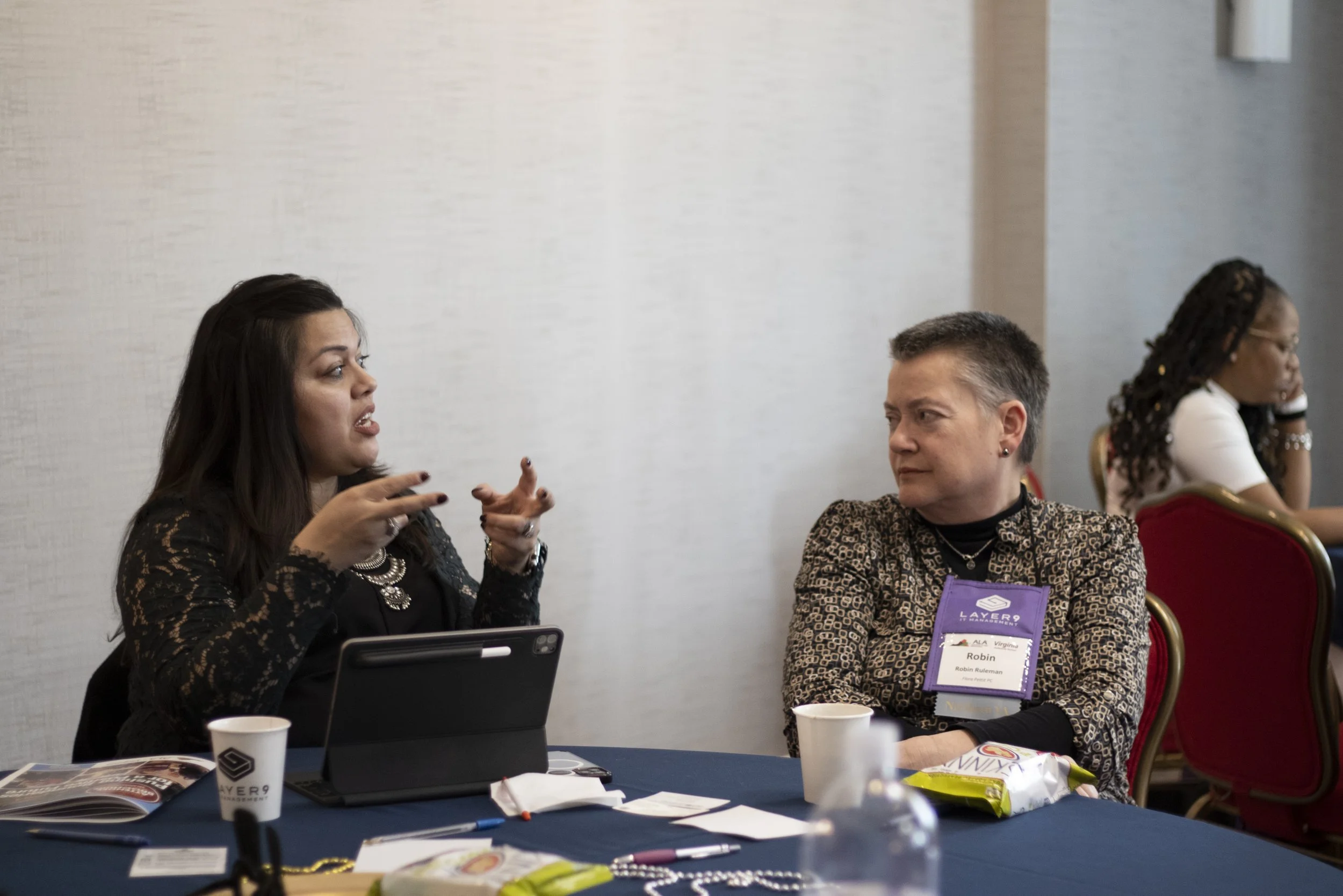 Three women sitting at a conference table engaged in a discussion at a professional event. The woman on the left is speaking, gesturing with her hands, while the woman on the right listens, wearing a nametag labeled Robin. The third woman is in the b