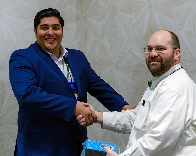 Two men shaking hands, one in a blue suit and the other in a white medical coat, smiling in a professional setting.