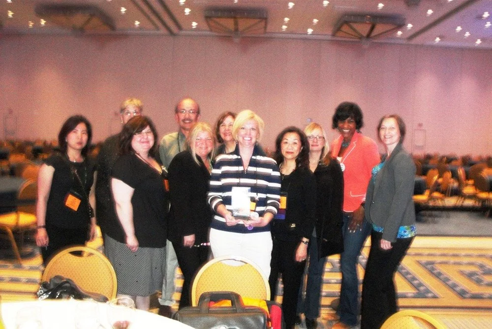 Group of ten women and one man standing together in a conference hall, with one woman in the middle holding an award, smiling at the camera.