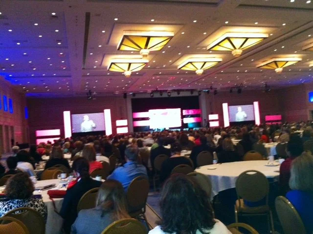 Large conference room filled with attendees seated at round tables, facing a stage with large screens displaying a presenter.