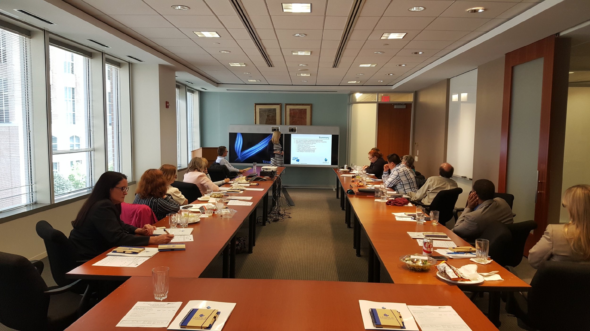A conference room with a long table surrounded by people listening to a woman presenting at the front. The room has large windows on one side and a projector screen displaying a presentation.