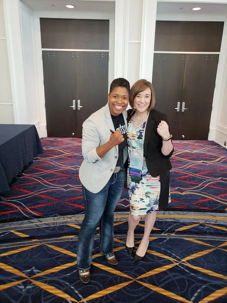 Two women standing on a patterned carpet in a conference room, smiling at the camera, with both raising one fist in a celebratory gesture.