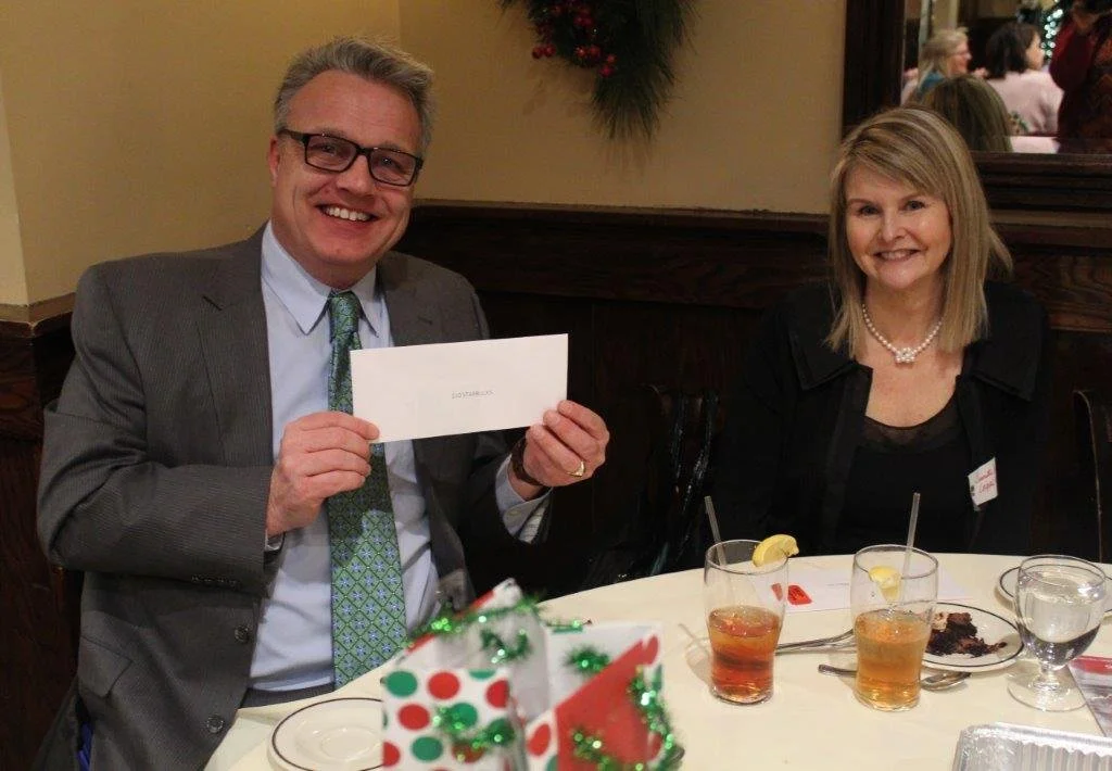 A man and woman sitting at a decorated table during a holiday celebration, smiling at the camera, with drinks, a dessert plate, and holiday decor visible.