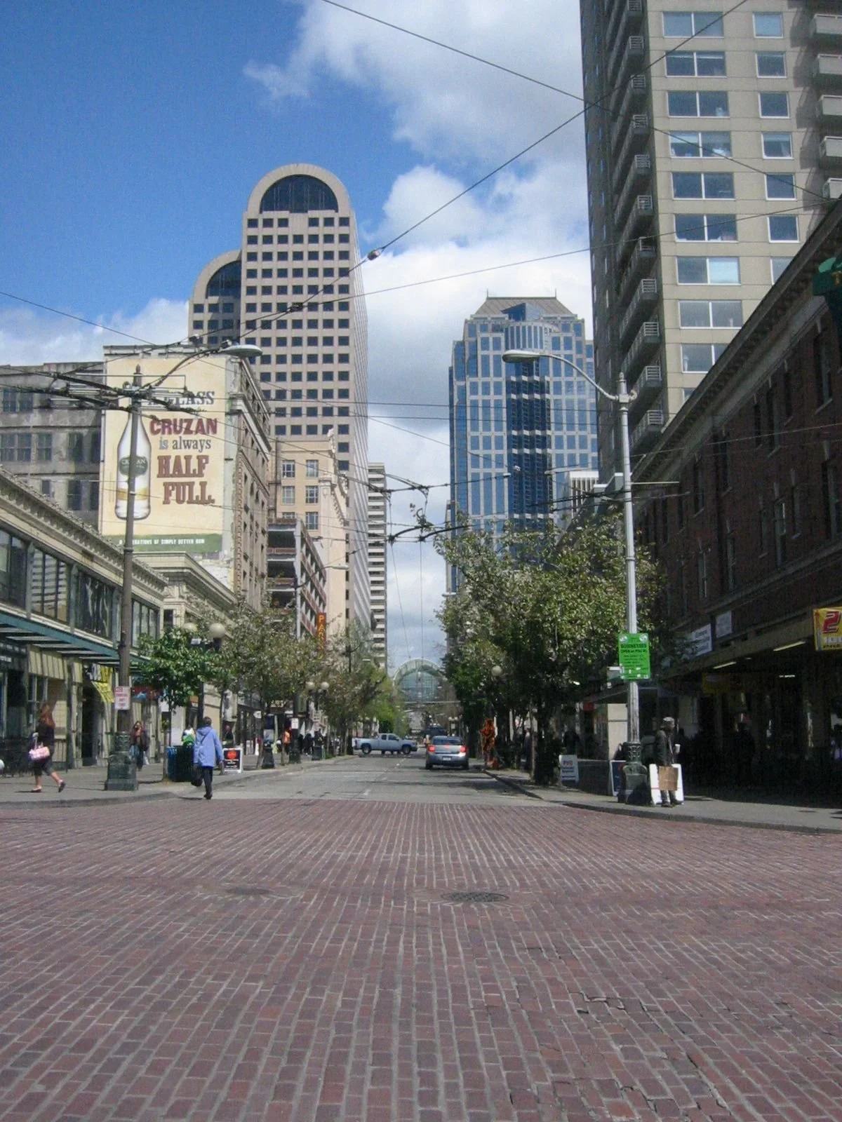 A city street with tall buildings, trees, pedestrians, and parked cars. Overhead wires cross the street, and a bridge is visible in the background. The sky is partly cloudy.