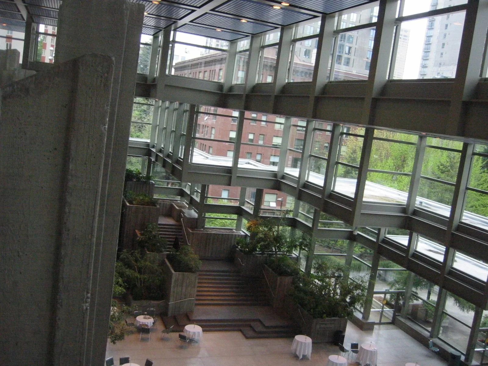 Interior view of a building with large glass windows, multiple floors, plants, and round tables with chairs, showcasing modern architecture.