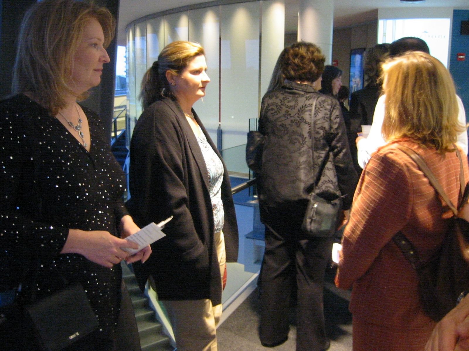 A group of women are standing in a line indoors, possibly waiting or attending an event. They are dressed in semi-formal attire, and some are holding papers or pamphlets.