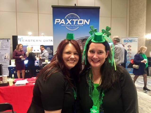 Two women wearing green hats and beads, celebrating St. Patrick's Day at a conference or expo, with booths and other attendees in the background.
