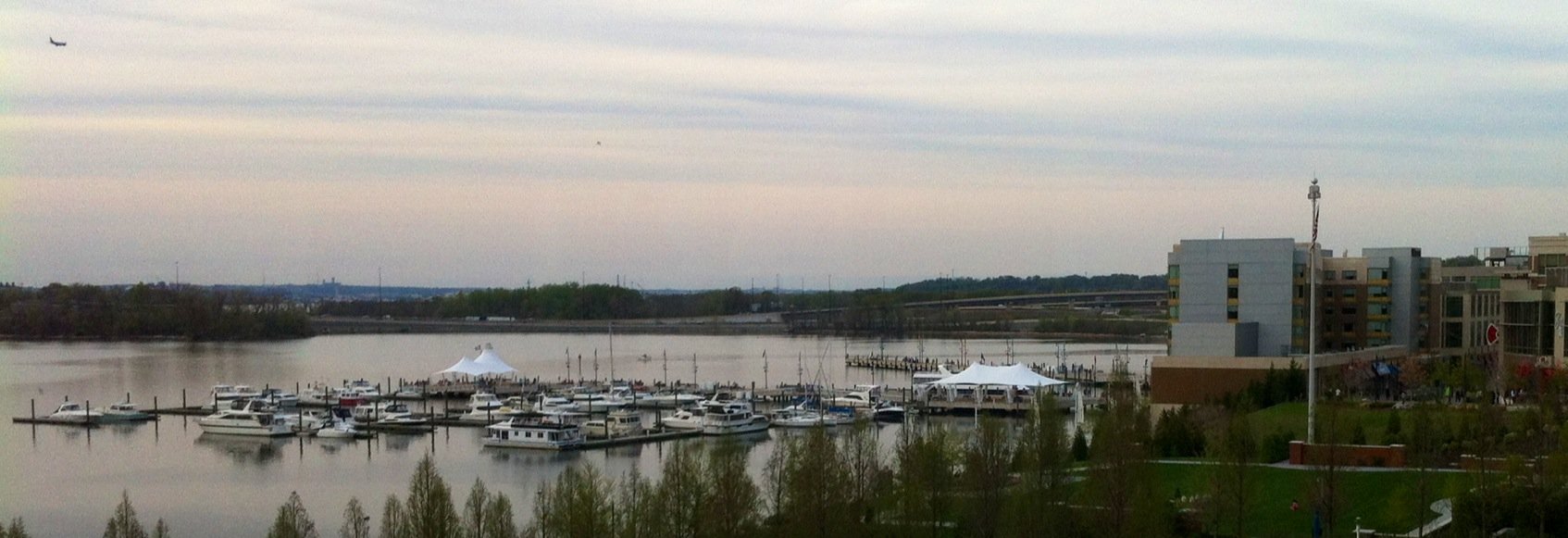 A marina with numerous boats docked on calm water, white tents on docks, modern apartment buildings on the right, a flagpole, and a cloudy sky.