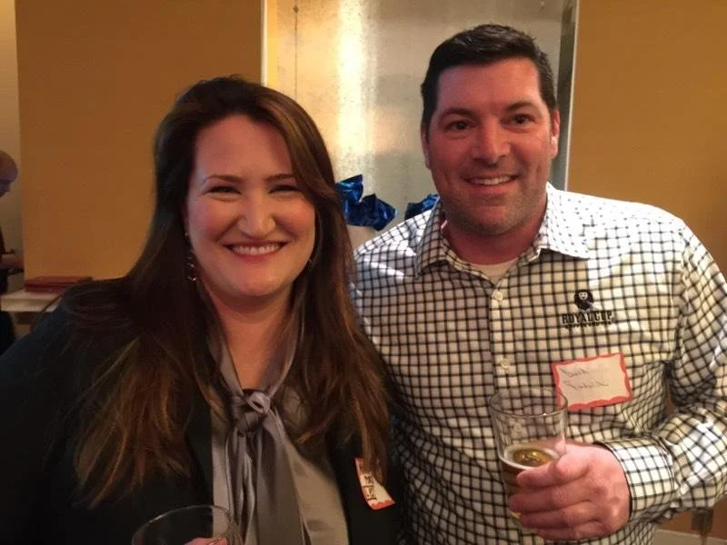 A woman and a man smiling and posing at a social event, holding drinks, with a warm indoor setting and a yellow wall in the background.