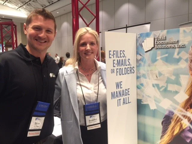 Two smiling people, a man and a woman, standing next to a display booth at a conference or expo. The man wears a black polo shirt and the woman is in a light blazer. They are both wearing name tags. The booth features a sign that says, 'E-FILES, E-MA