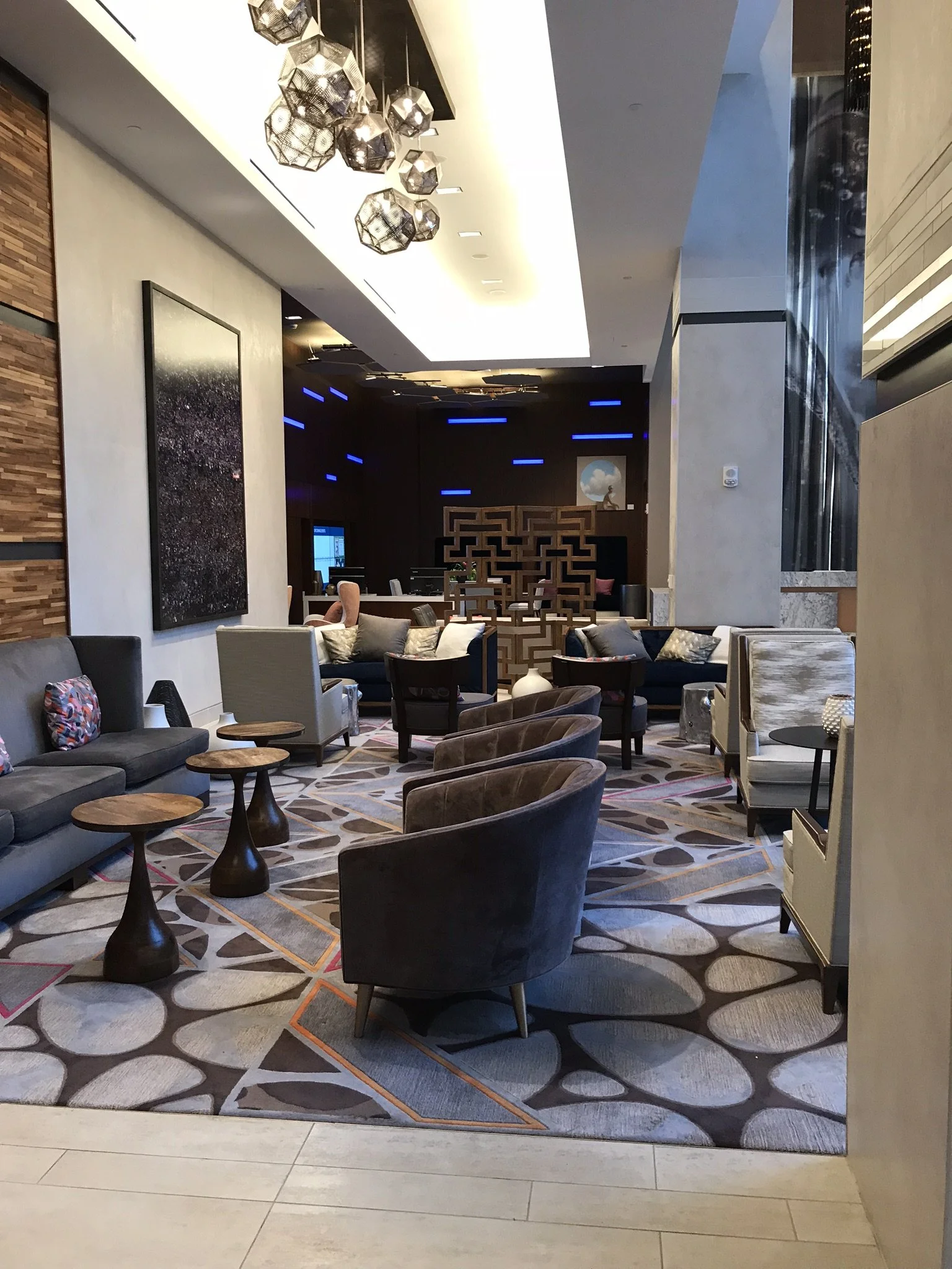 A modern hotel lobby with seating areas, a patterned carpet, decorative lighting fixtures, wall art, and a backlit feature wall with blue lights.