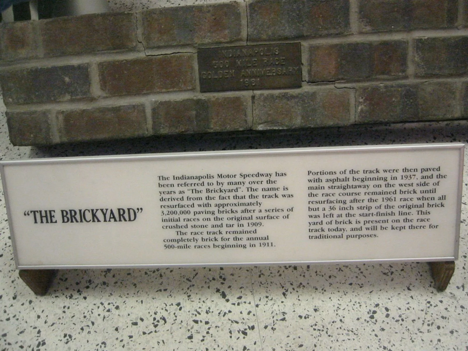 Brick wall with a plaque and an informational sign about the Indianapolis Motor Speedway and the origin of the name "The Brickyard."