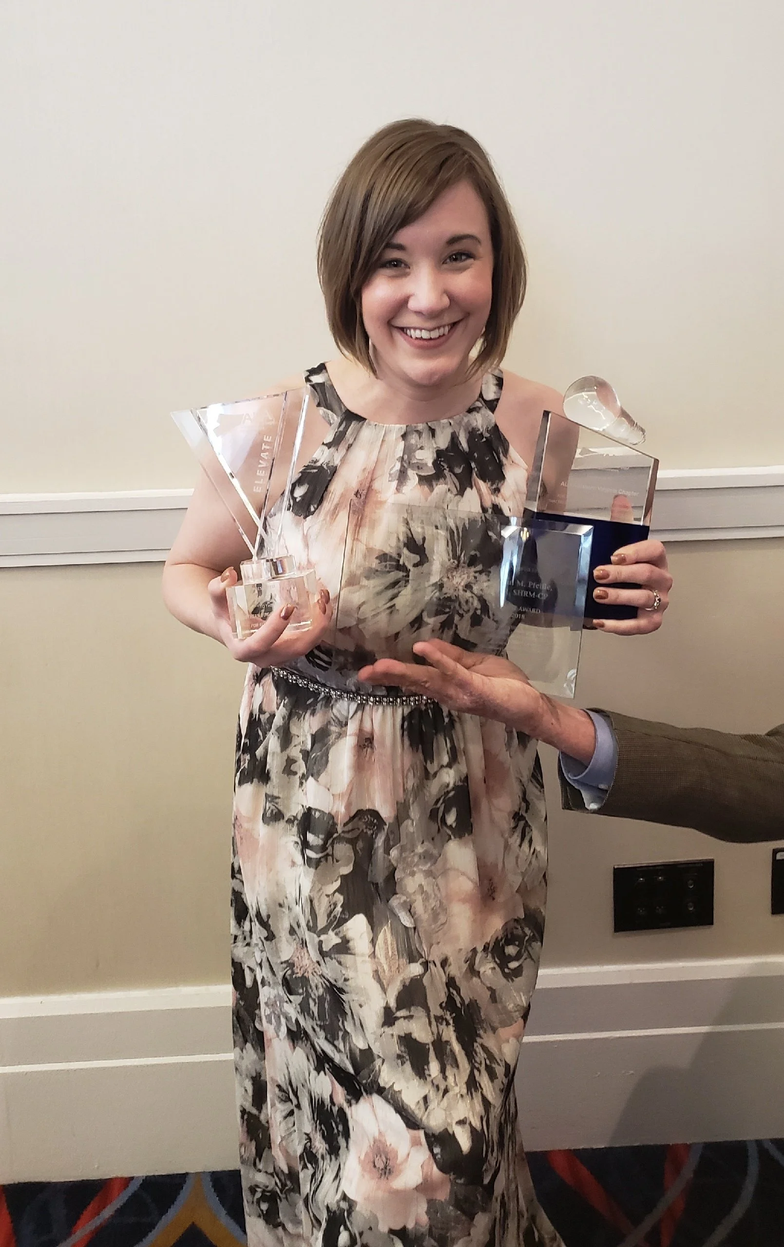 A woman smiling and holding two awards, a crystal triangle and a glass sculpture, in front of a plain wall at an indoor event.