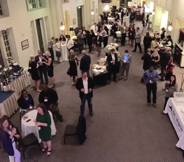 People attending a networking event or conference in a large, well-lit room with tables set up for food and drinks, some engaging in conversations, others browsing or standing around.