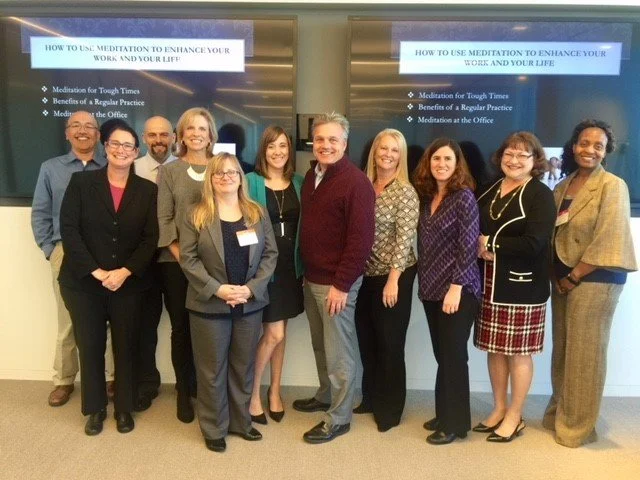 Group of ten people standing in front of two screens displaying a presentation about using meditation to enhance work and life. The group is smiling and dressed in business casual attire.