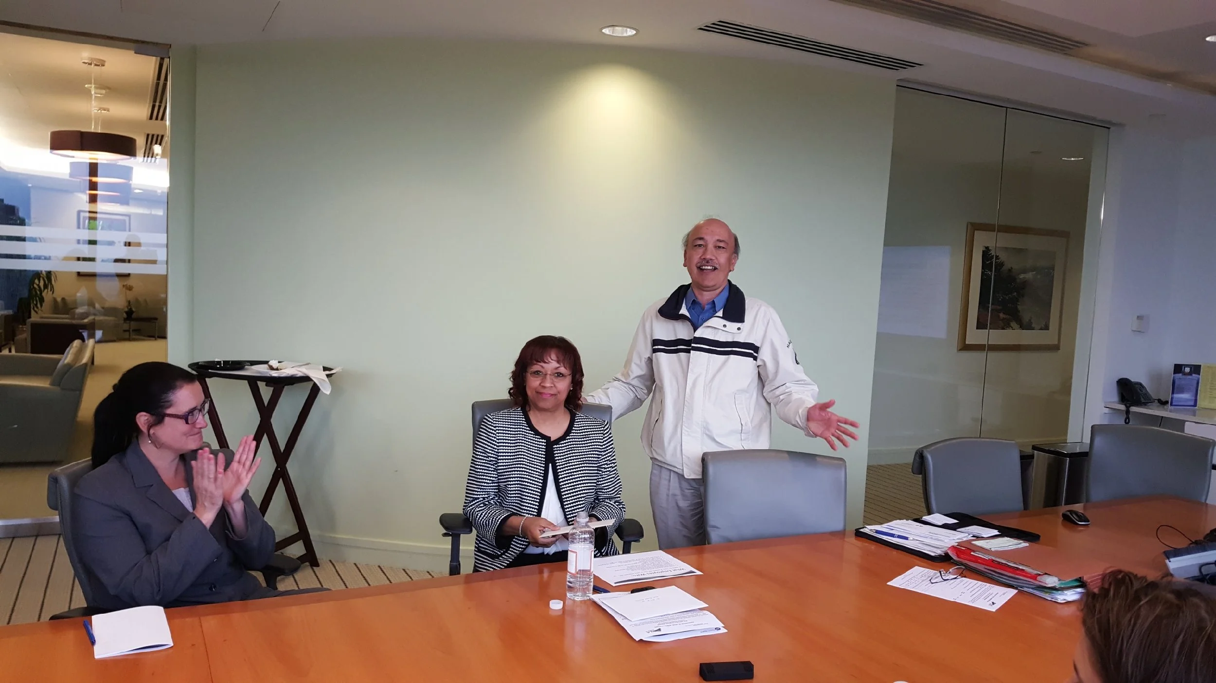 People in a meeting room, one woman clapping, one woman sitting at the table, and a man standing with arms open.
