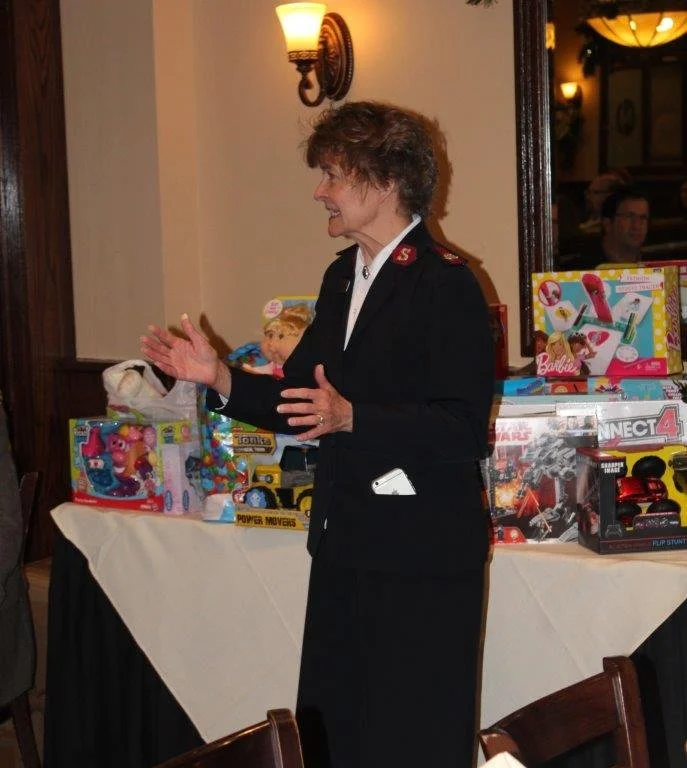 A woman in a military or formal uniform talking and gesturing with her hands at a table with toys in the background.