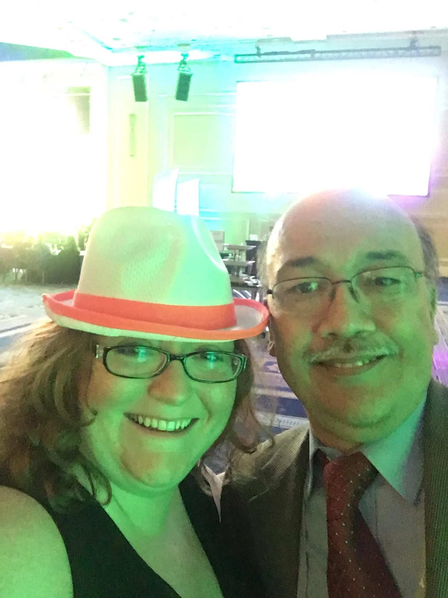 Two people taking a selfie in a brightly lit room with a large screen, speakers, and chairs. One person is wearing glasses and a black top, while the other person is wearing a sparkly red dress and a colorful hat.