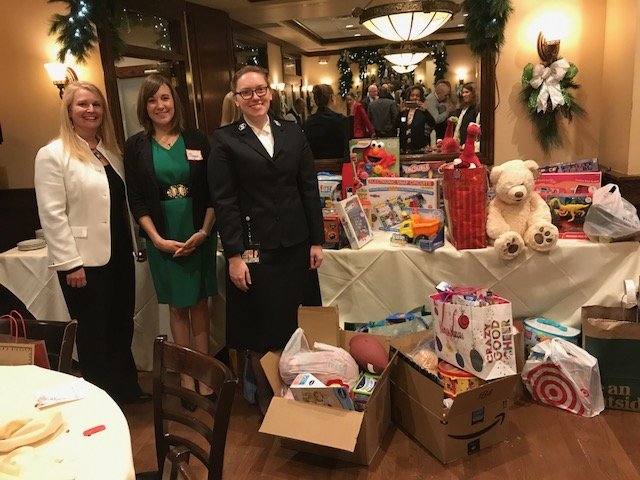 Three women stand together in a festive room with holiday decorations, smiling in front of a table filled with toys, including Elmo, a teddy bear, and various boxed gifts, as well as cardboard boxes and gift bags.