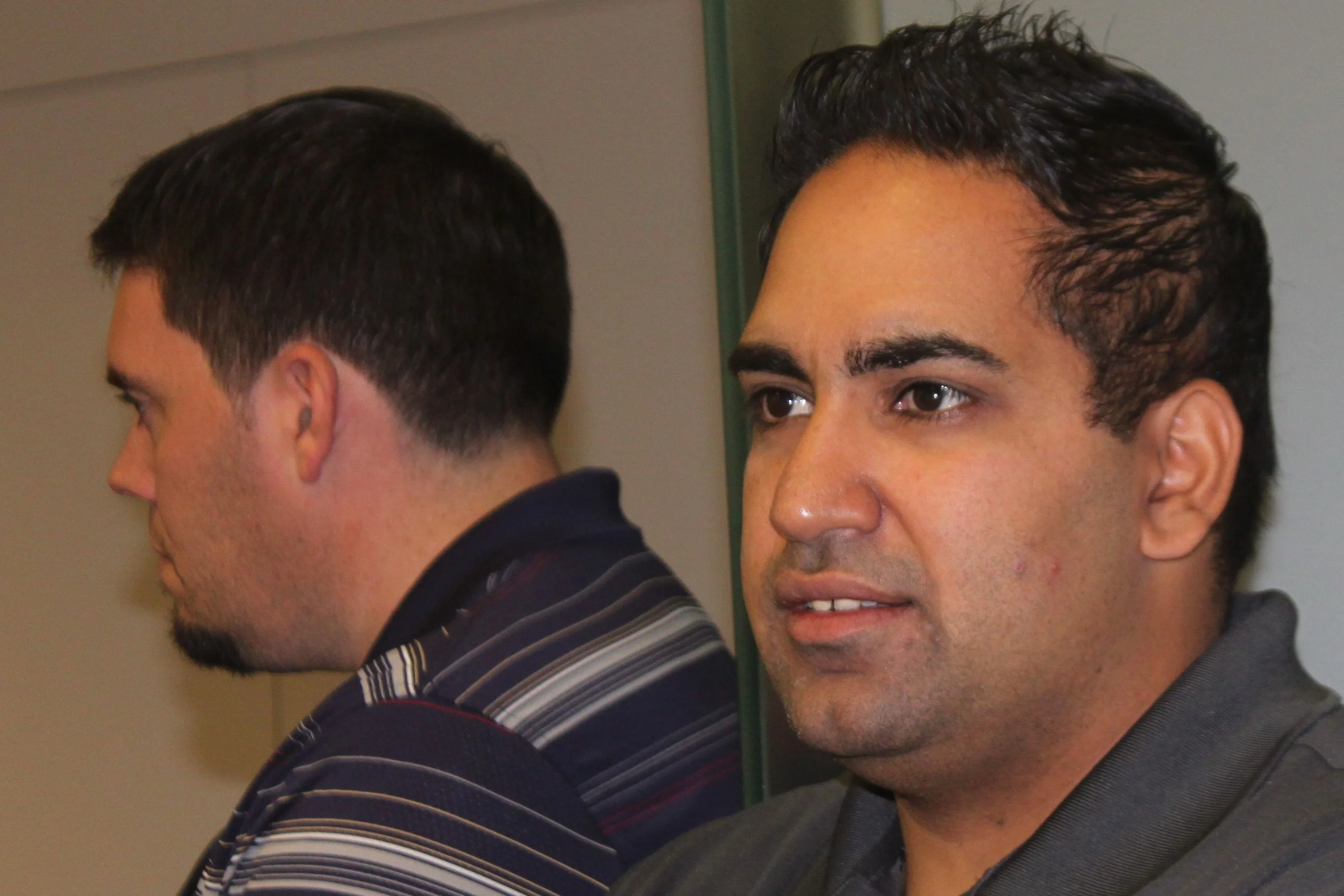 Two men's faces in profile, one facing forward and the other facing sideways, with dark hair and wearing striped shirts.