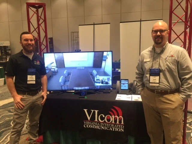 Two men standing next to a display table with a large screen TV at a conference or expo. The table has a black tablecloth with the logo 'Vlcom Virginia Integrated Communication.' Both men are smiling, wearing conference name tags, and business-casual