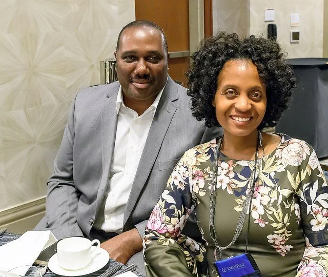A man in a gray suit and a woman with curly hair and a floral dress sitting at a table with cups and papers, smiling at the camera in a conference room.