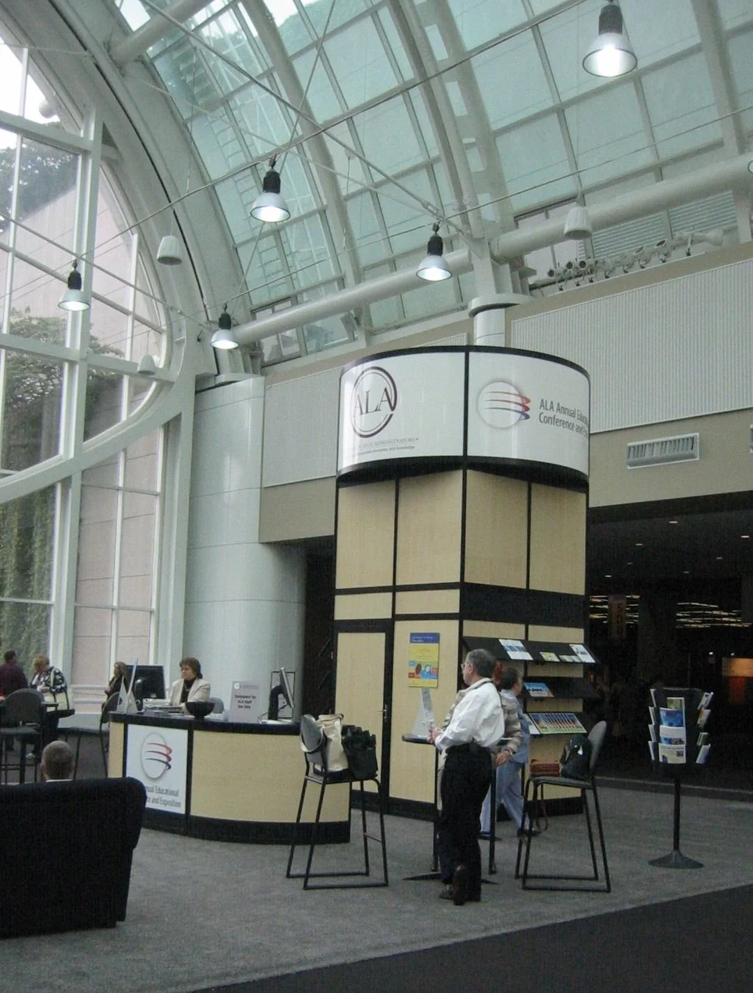 Interior of a modern conference center with people at registration desks, digital screens, and signs for the American Library Association annual event.