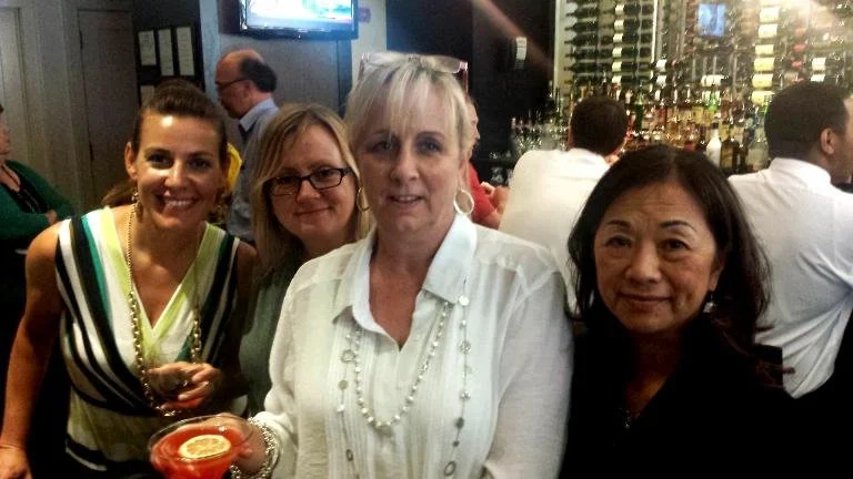Four women posing for a photo in a bar or restaurant, with wine bottles and patrons in the background.