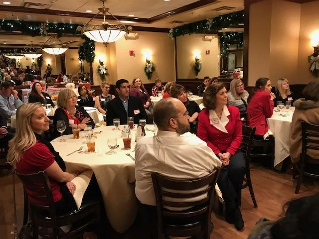 People in a banquet hall attending a formal event or gathering. Attendees are seated at round tables, listening to a speaker or presentation. The room is decorated with lights and floral arrangements.