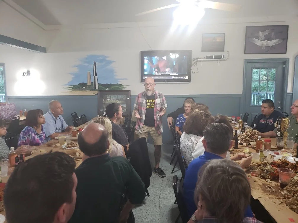 A group of people gathered around a long dining table in a room with artwork on the walls, listening to a man standing and speaking to the group.