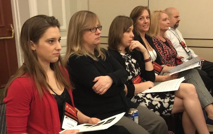 Six women sitting in a row at a conference or seminar, holding papers or programs, attentively listening to a speaker.