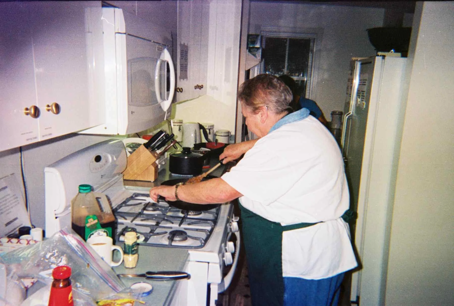 An elderly woman cooking on a stove in a small kitchen, with various kitchen utensils and food items on the counter.