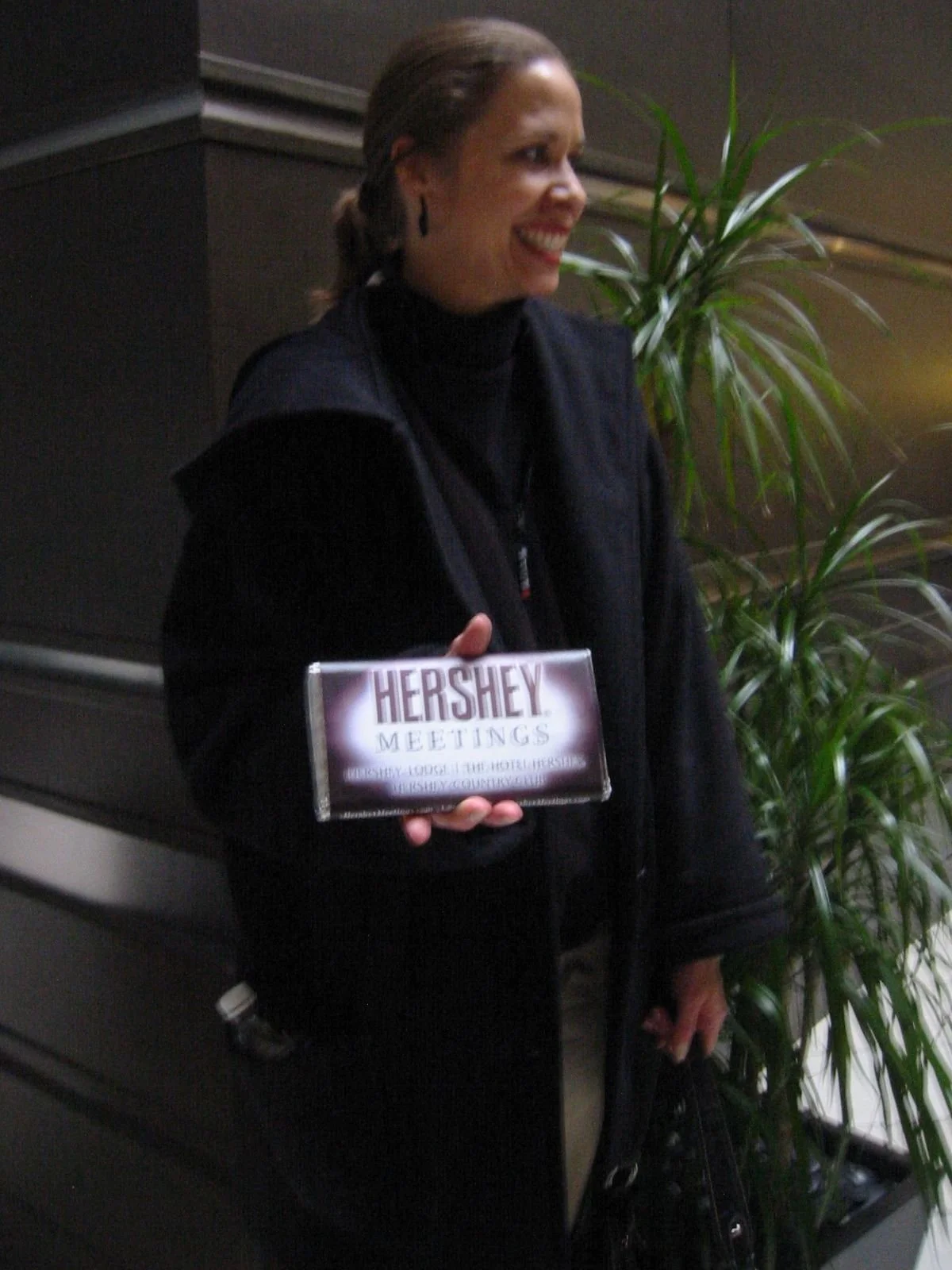 A woman smiling and holding a Hershey's Meetings box, standing indoors near a plant and dark wood paneling.