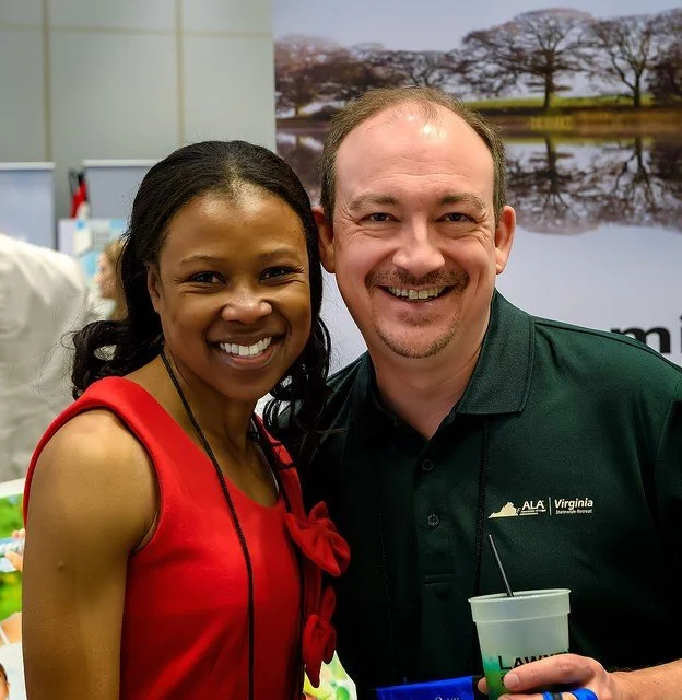 Two people smiling, a woman in a red top and a man in a dark green polo with a Virginia logo, in an indoor setting with a nature picture in the background.