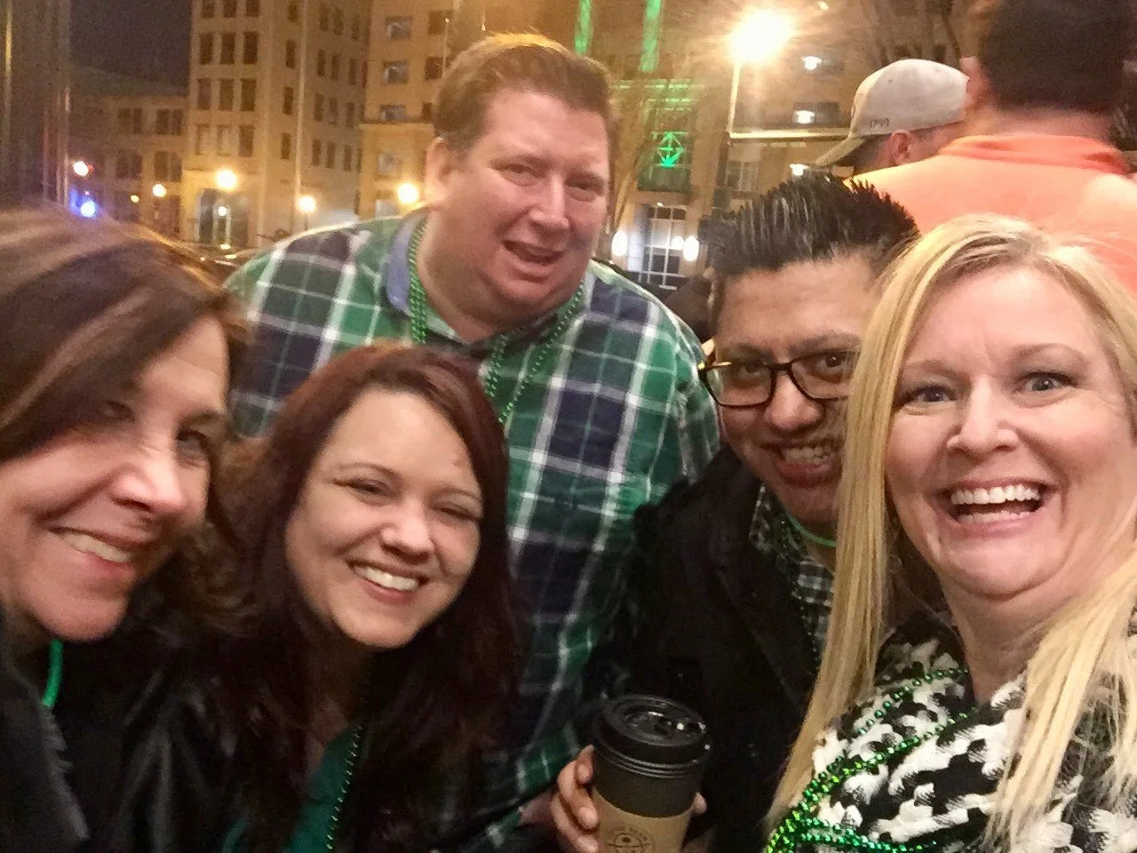 Group of five friends smiling and posing for a photo outside at night during a celebration or Mardi Gras event with some wearing green beads, in an urban setting with buildings and streetlights in the background.