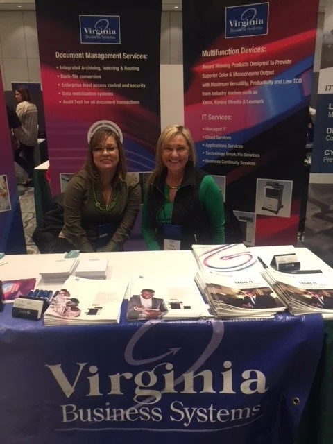 Two women sit behind a table at a Virginia Business Systems booth, with promotional banners and brochures displayed.
