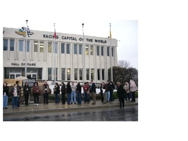 Group of people standing in line outside the National Speedway Museum, known as the Hall of Fame, in front of a large building with a sign reading 'Racing Capital of the World'.