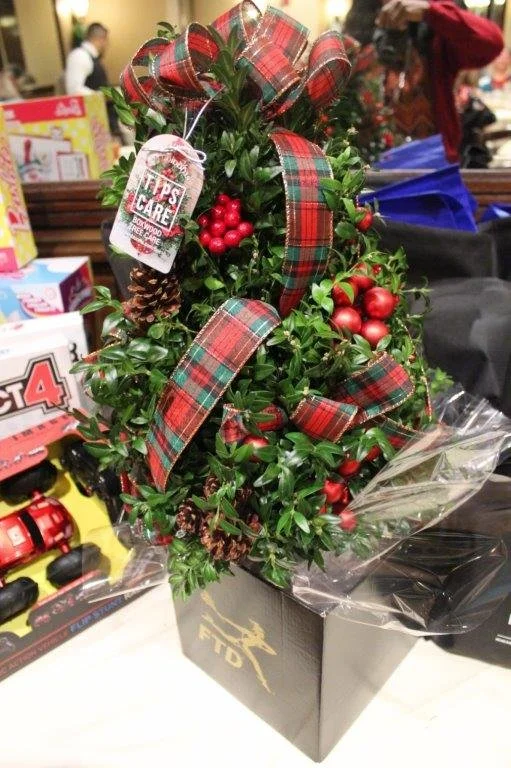 A small decorative Christmas tree with red berries, pinecones, and plaid ribbons, wrapped in clear plastic packaging and placed in a black box